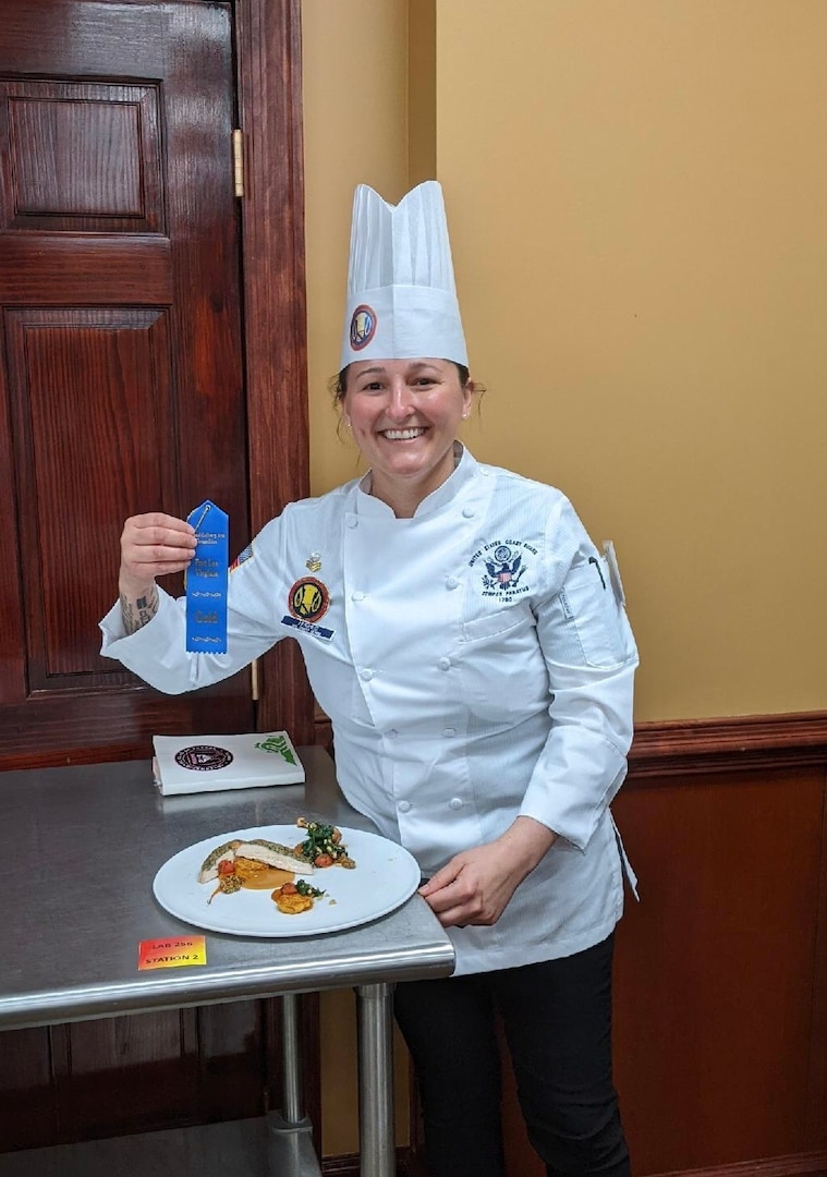 Petty Officer 1st Class Danielle Hughes stands in front of her winning dish holding her gold medal ribbon after placing first in the category of Military Master Chef of the Year, March 10, 2022. USCG photo.