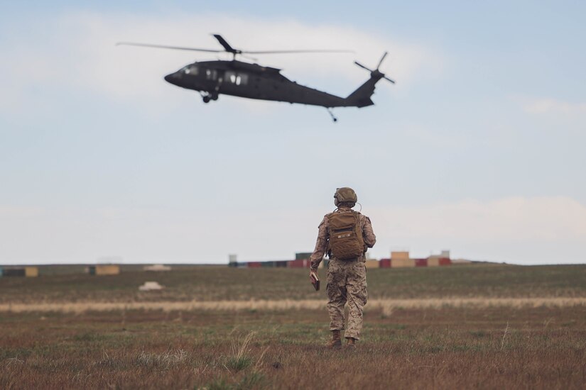 A Marine walks through a field as a helicopter flies above.