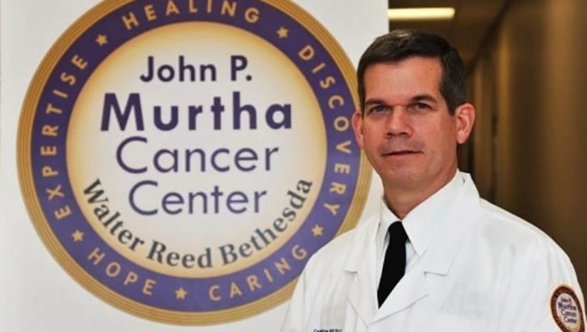 A man in a lab coat stands next to a large logo on a wall.