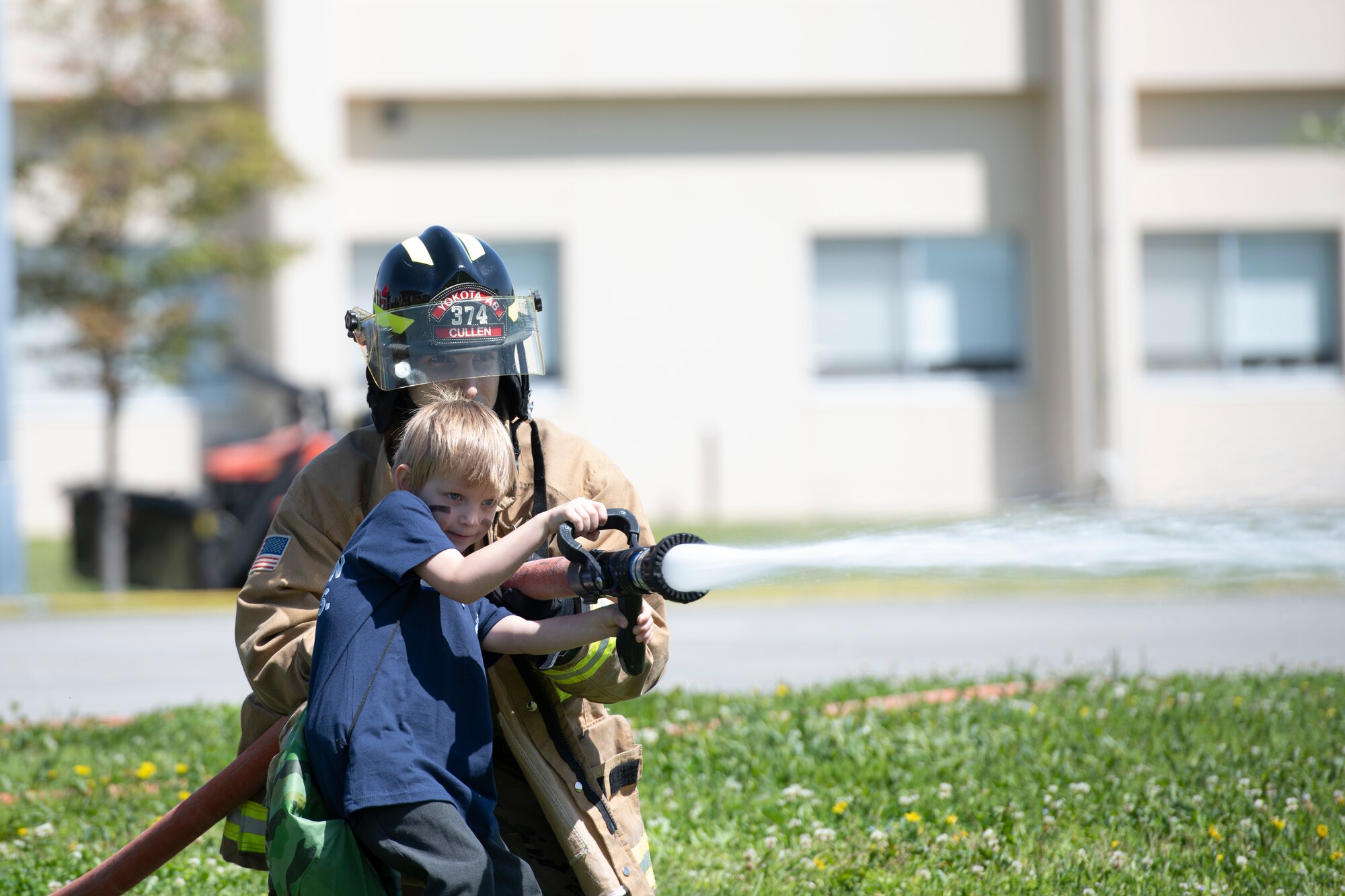 Senior Airman Peter Cullen, 374th Civil Engineer Squadron fire protection journeyman, back, assists Landon Crawford, Operation Kids Understanding Deployment Operations participant, with a fire hose during Operation K.U.D.O.S. at Yokota Air Base, Japan, April 30, 2022. Operation K.U.D.O.S. consisted of pre-deployment processing and mock deployment scenario to help children understand what it is like for their parent to deploy. (U.S. Air Force photo by Tech. Sgt. Joshua Edwards)