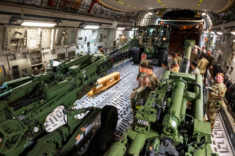 Uniformed service members stand near artillery inside the cargo loading area of an airplane.