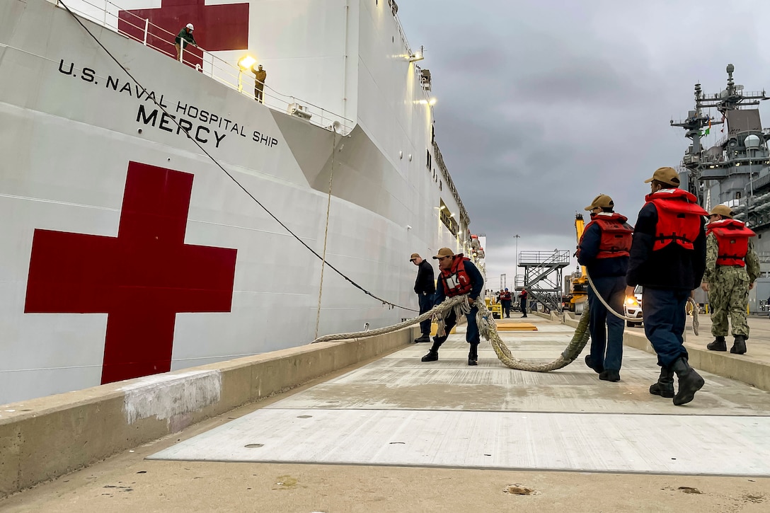 A group of sailors on a deck pull ropes to undock a large ship.