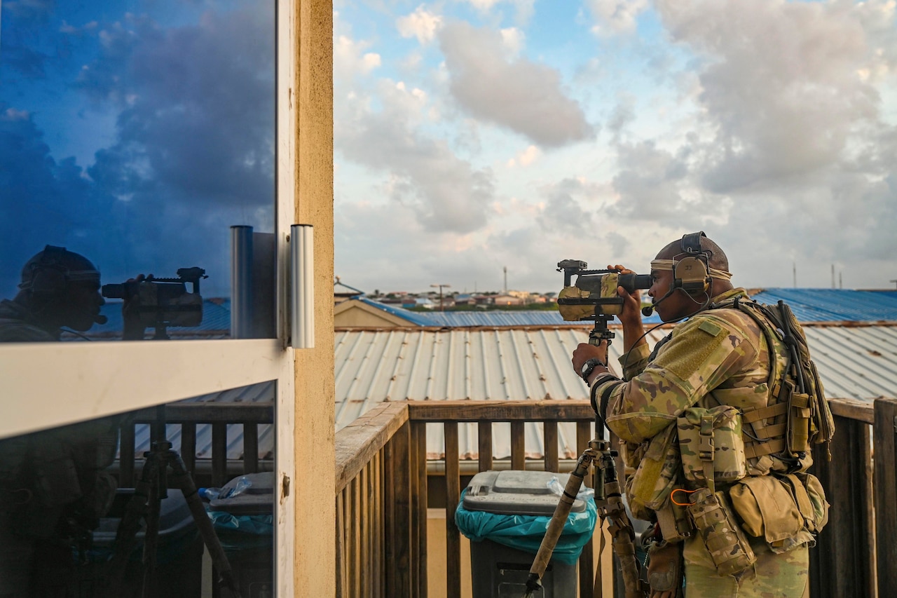 A soldier looks out of binoculars from a tower.