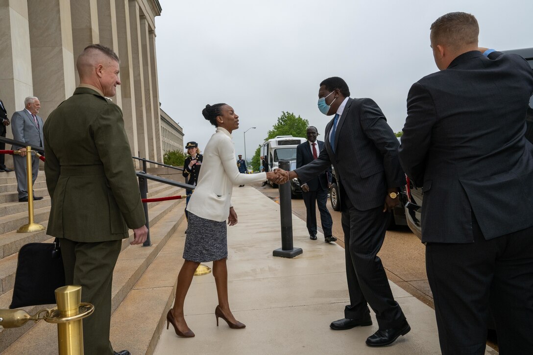 A woman and a man wearing a face mask shake hands in a group of people at the bottom of the steps outside the Pentagon.