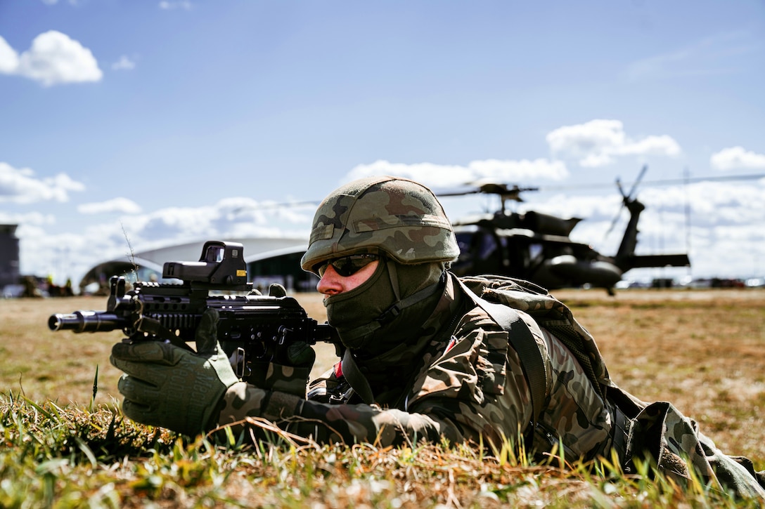 A Polish soldier holds a rifle in a prone position on the ground.