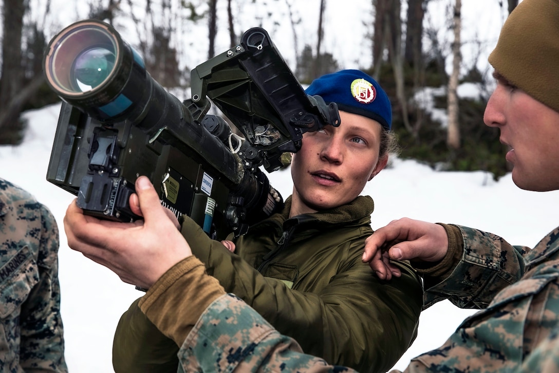 A uniformed woman holds a shoulder-fired missile launcher while a Marine assists at her side.