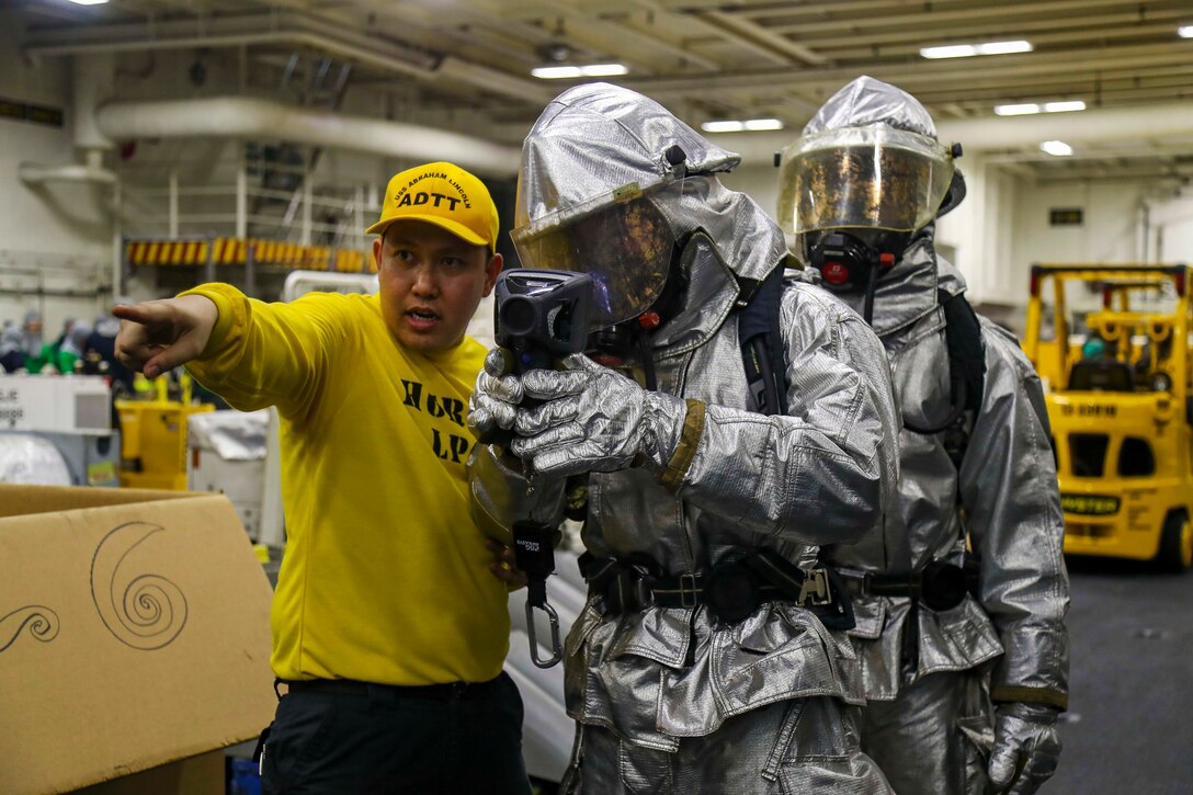 A sailor stands next to and instructs two others in hazmat suits.