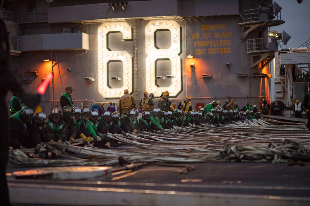 Sailors observe a long line of fellow sailors kneel to pull flat hoses on a ship's deck.