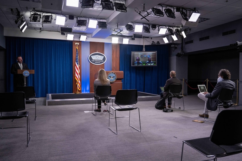 Two service members brief a room of people from a television screen.