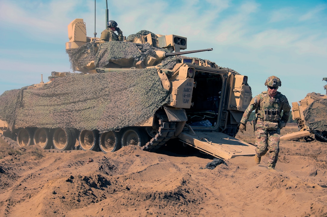 One soldier sits in the top of a tank while another soldier walks down a hill of sand.