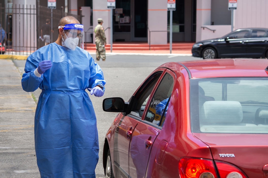 A soldier wearing personal protective equipment holds a nasal swab while standing near the open driver-side window of a car.