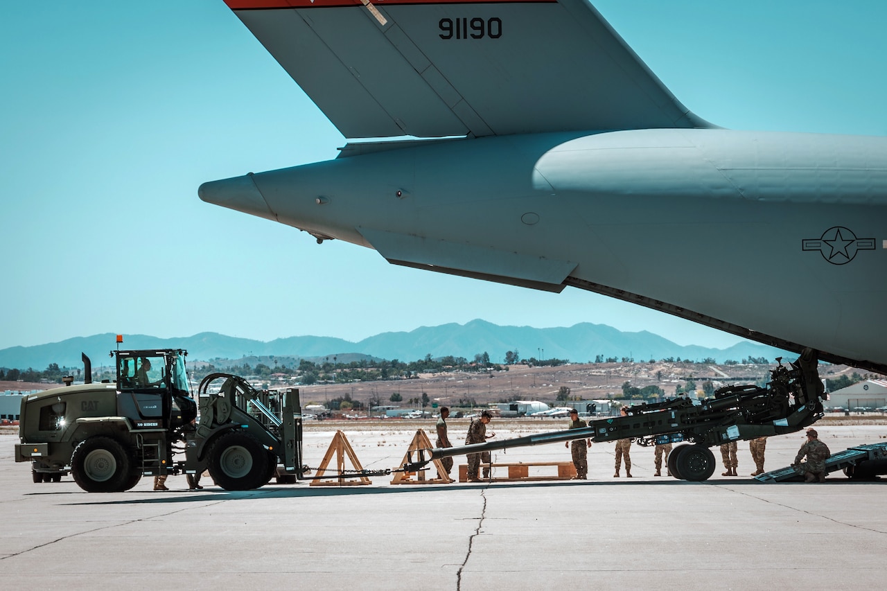 Troops tow a large artillery gun from a vehicle into the back of an airplane.