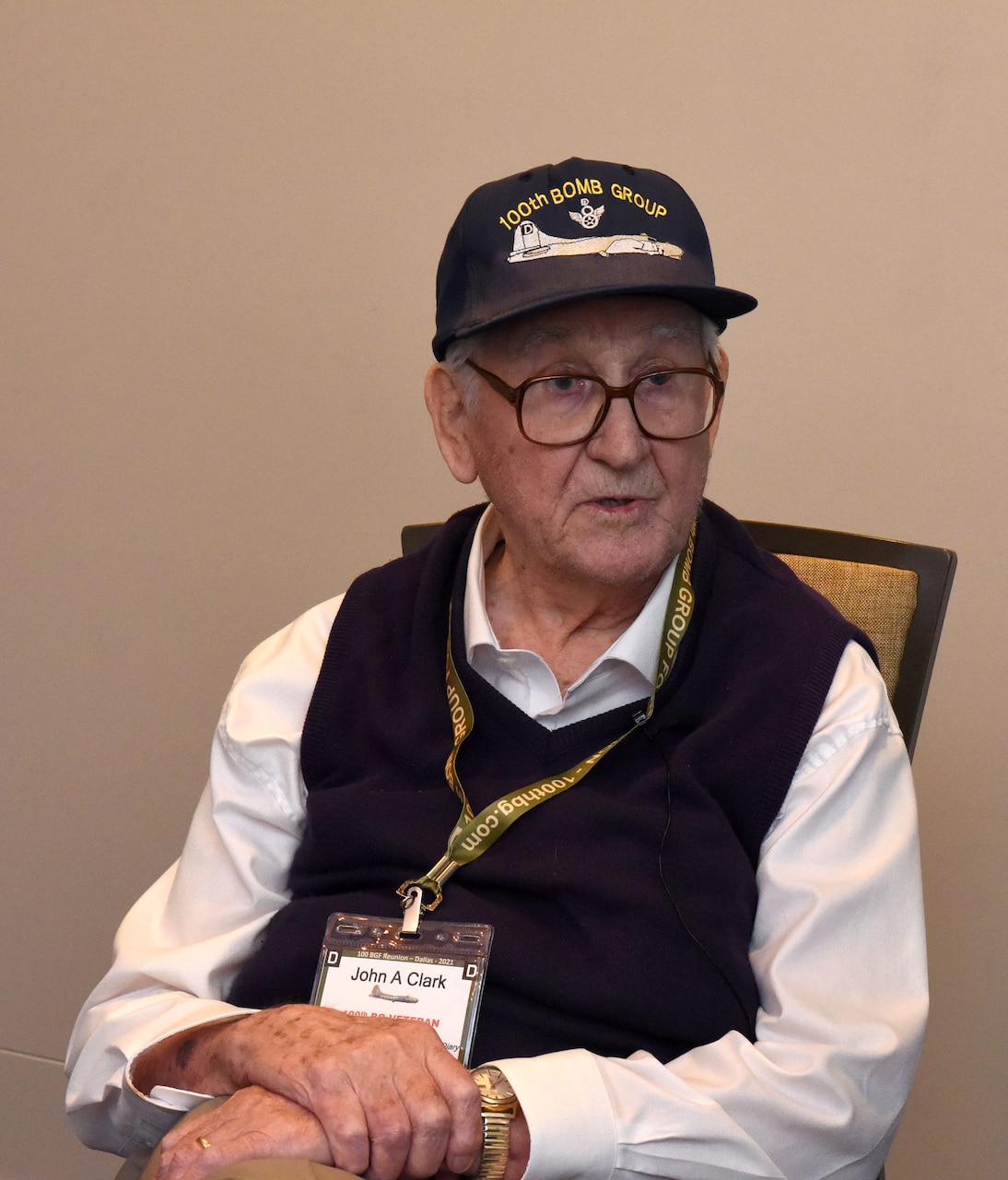 An elderly man wearing a cap that reads “100th Bomb Group” sits in a chair with his hands folded across his lap.