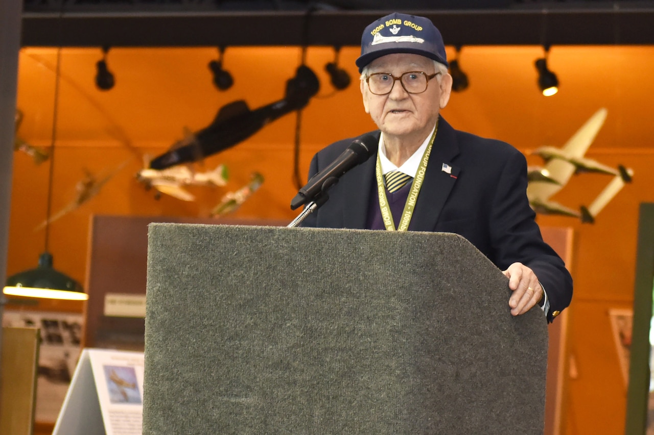 A man wearing a ball cap that reads “100th Bomb Group” stands at a podium.