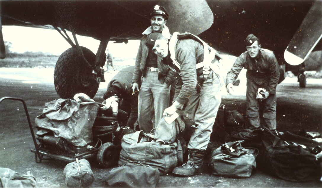Three service members stand among luggage beneath an aircraft, black-and-white photo.