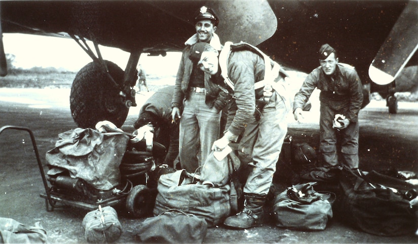 Three service members stand among luggage beneath an aircraft, black-and-white photo.