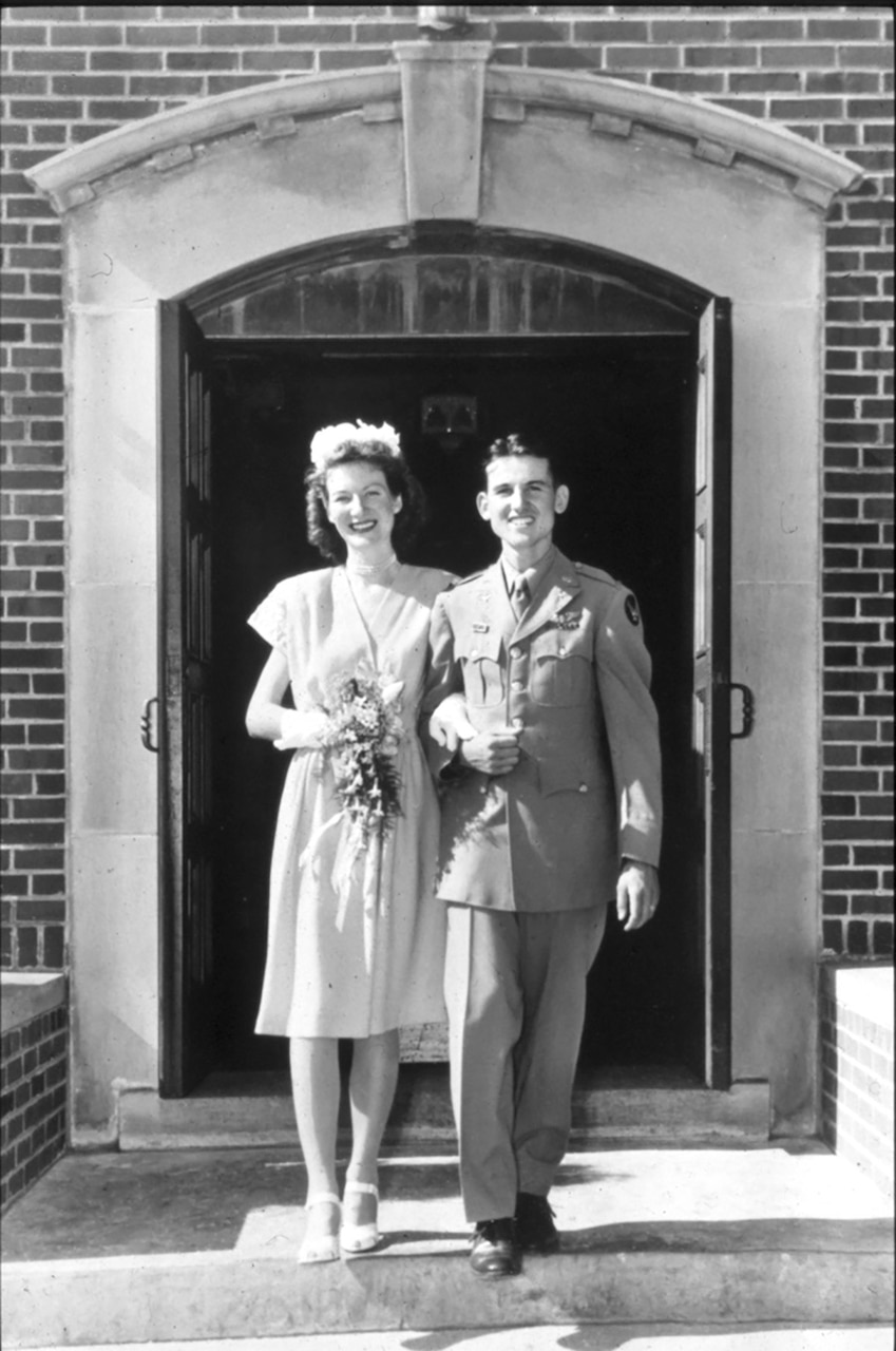 A bride and a groom wearing a military uniform stand at the top of steps and pose for a black-and-white photo.