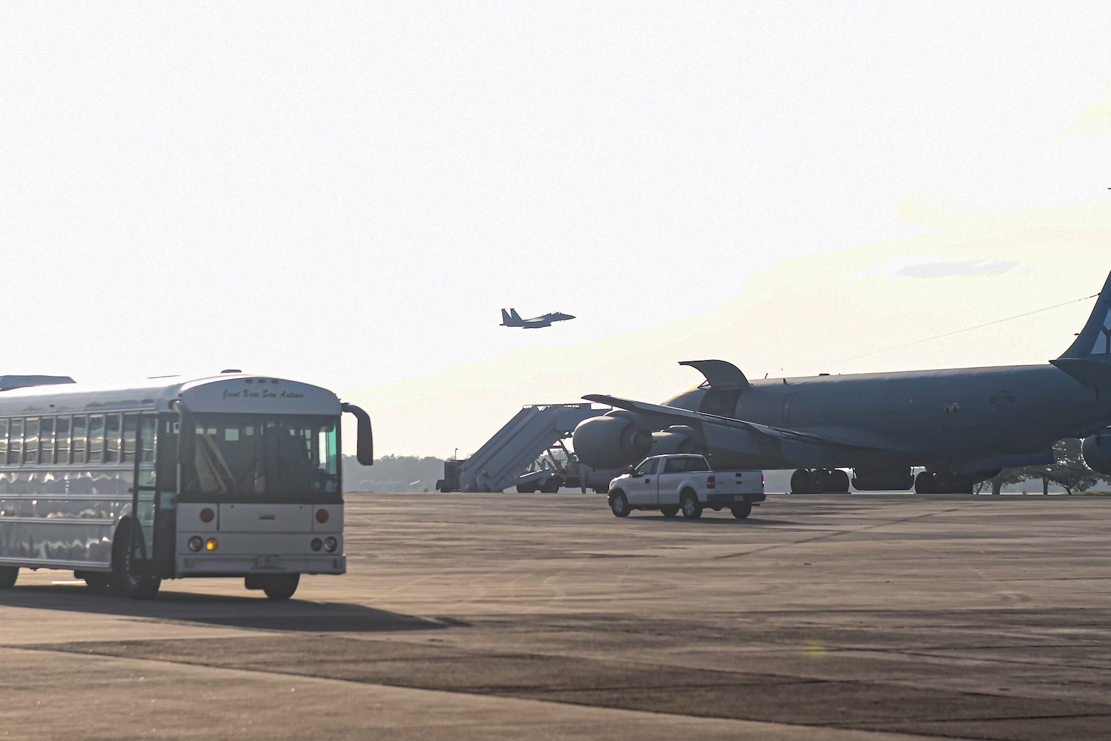 Fighter jet takes off over the horizon with a passenger bus and air refueling tanker on the airfield below.