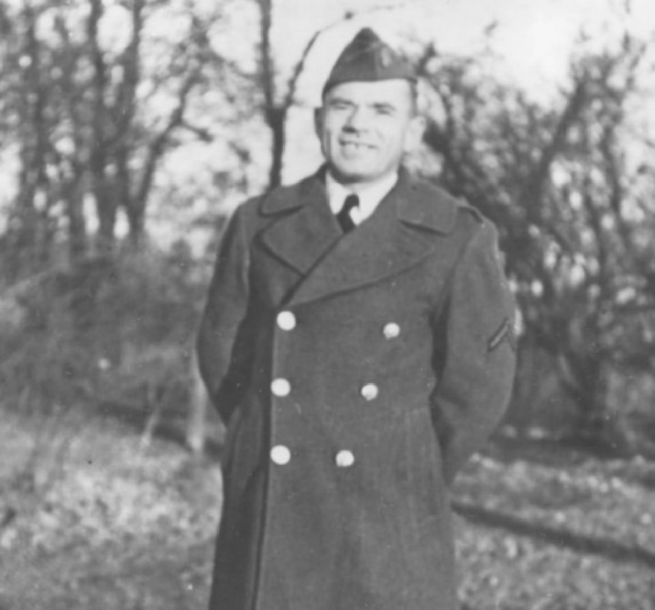 A man wearing a peacoat and military cap smiles with his hands behind his back.