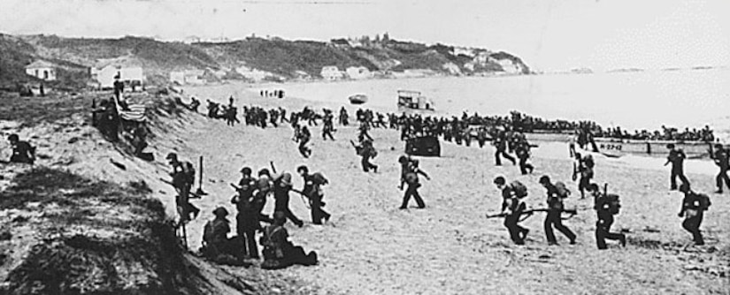 Hundreds of soldiers get off boats and walk toward bluffs on a beach.