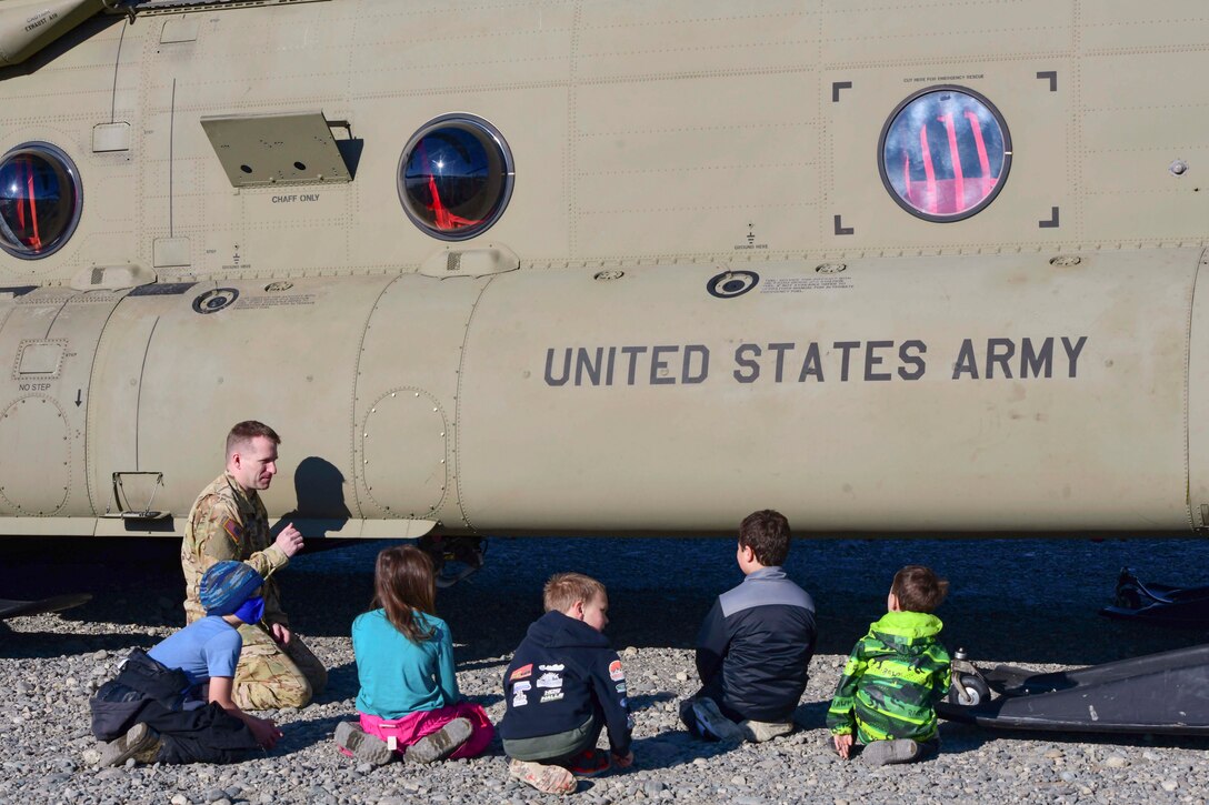 A soldier speaks to a group of children while kneeling next to a helicopter.
