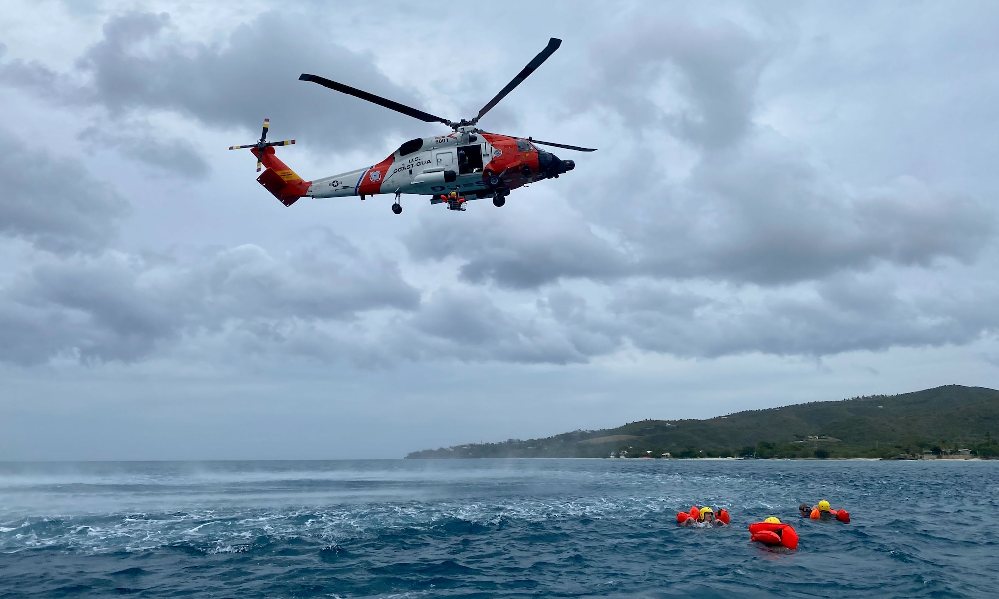 A helicopter hovers over water. Three people in life vests are in the water.