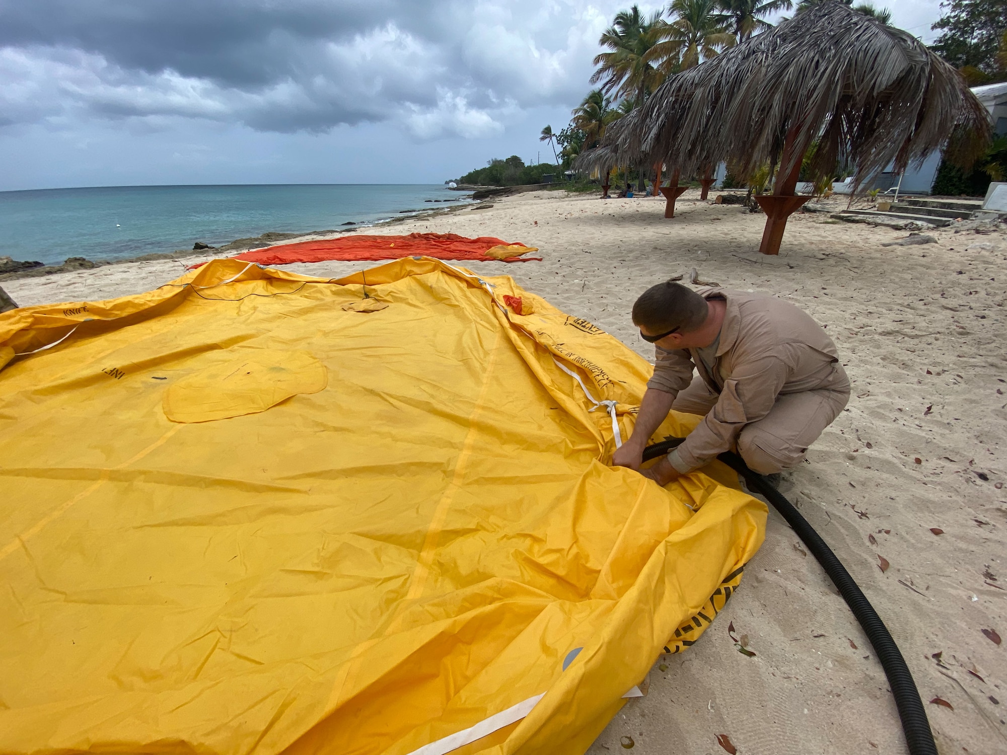 A large yellow raft lays on the beach. An Airman crouches to it, inflating it.