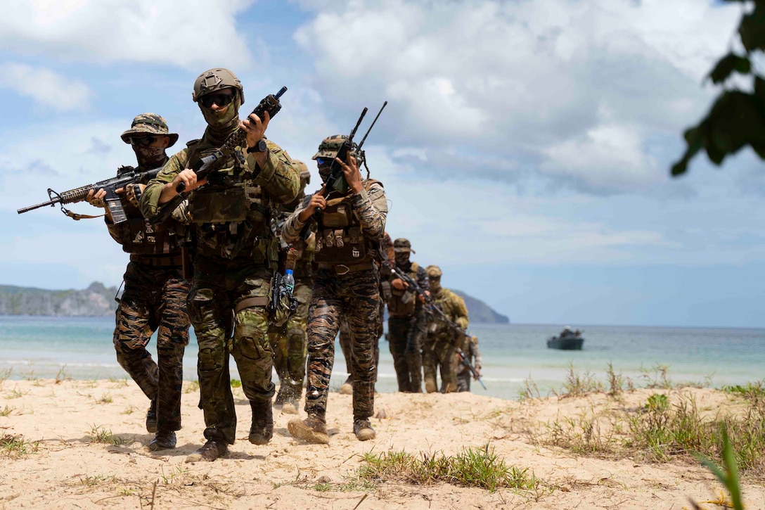 A group of service members  carrying weapons walk on a beach.