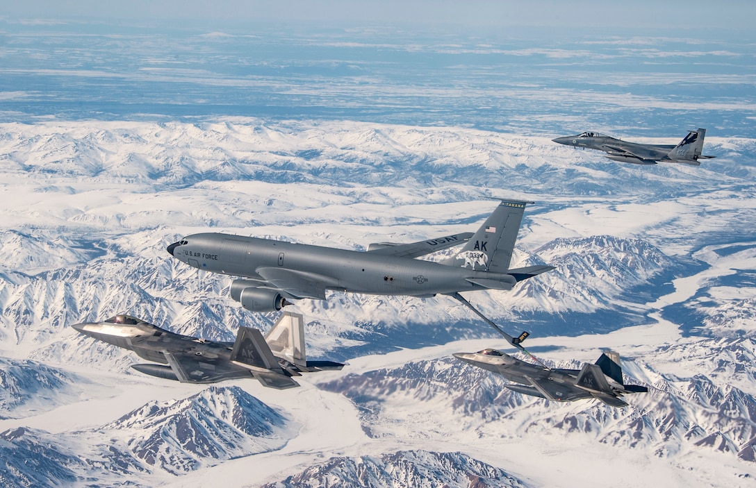 An aircraft fuels one of three others flying over snowy mountains.