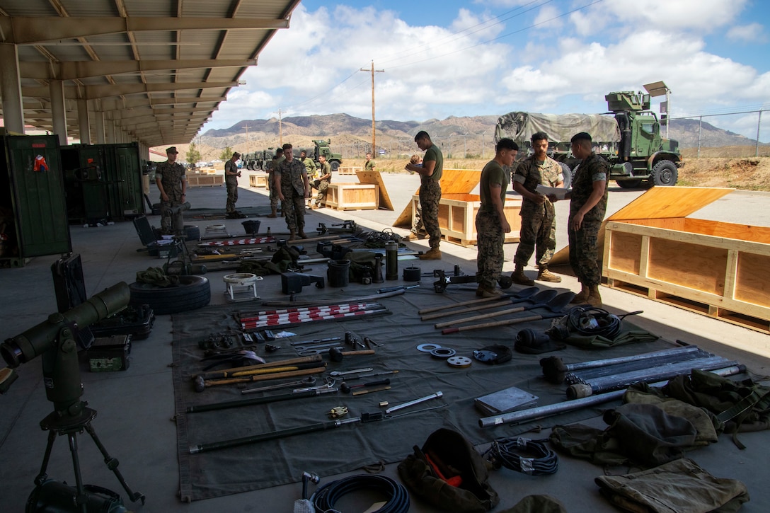 Service members stand among equipment that is laid neatly on the ground near open wooden crates.