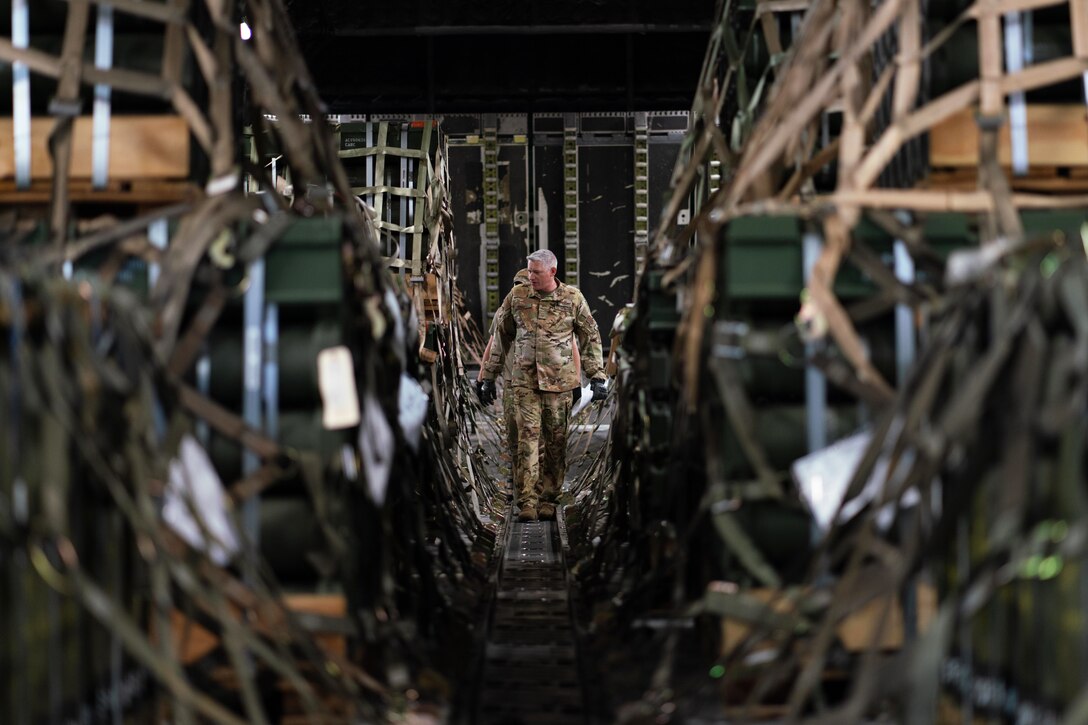 Two uniformed personnel walk between stacked pallets of supplies in the cargo hold of an aircraft.