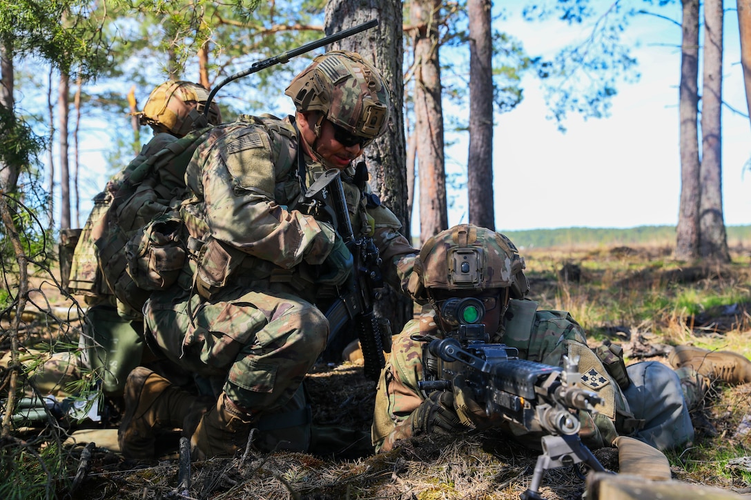 An armed soldier kneels and touches the shoulder of another soldier wielding a rifle in a prone position.