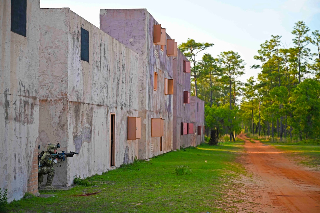 An airman holding a weapon kneels between two buildings.