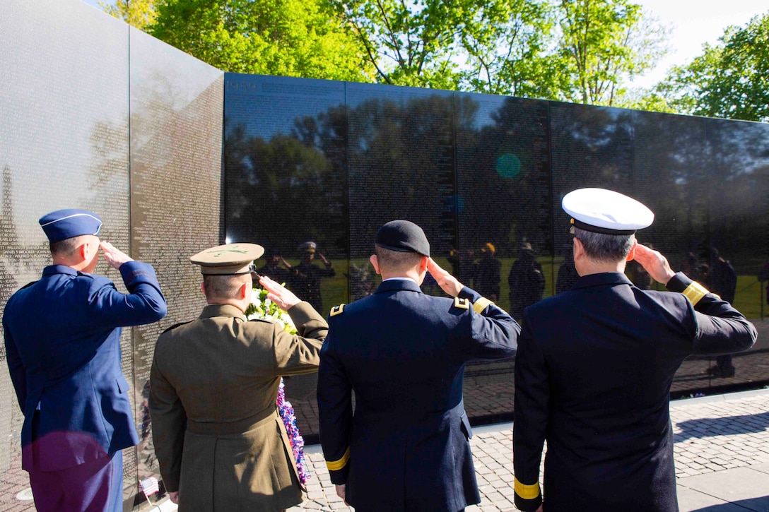 Four service members salute at a memorial wall.