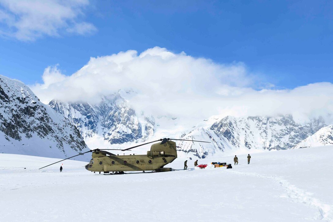 Soldiers offload equipment from a parked helicopter in a snowy area with a mountain in the background.