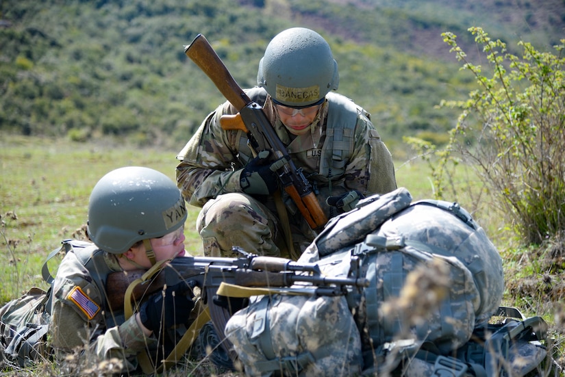 One service member in a prone position aims a rifle. Another service member is kneeling on the ground.