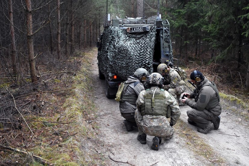 Four military personnel kneel on the ground, in the woods, behind a tactical vehicle.