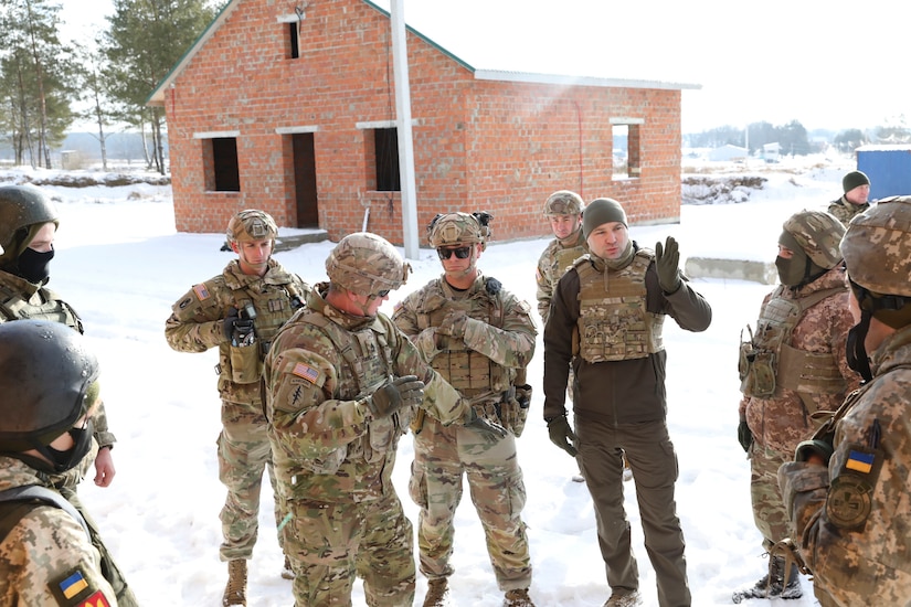 Military personnel stand together outdoors, on the snow-covered ground, outside a small brick house.