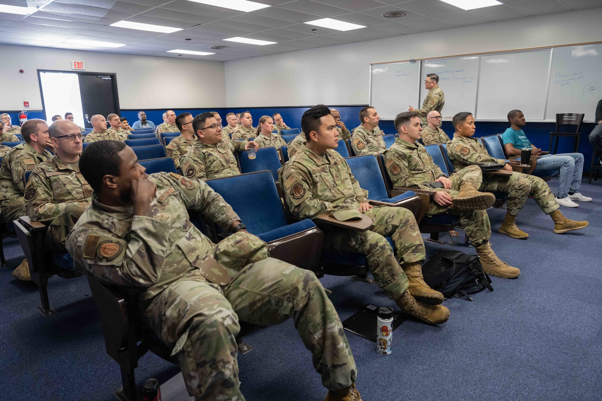 Military members sit in a room during a presentation.