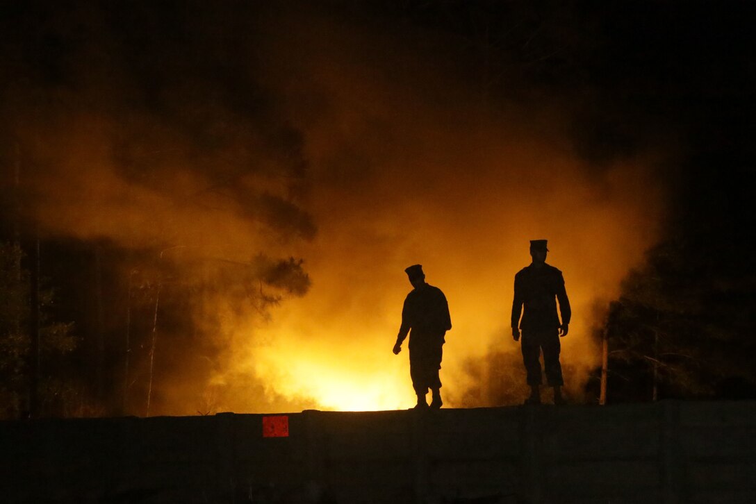 Marine Corps recruits stand next to each other as flames burn in the background as shown in silhouette.