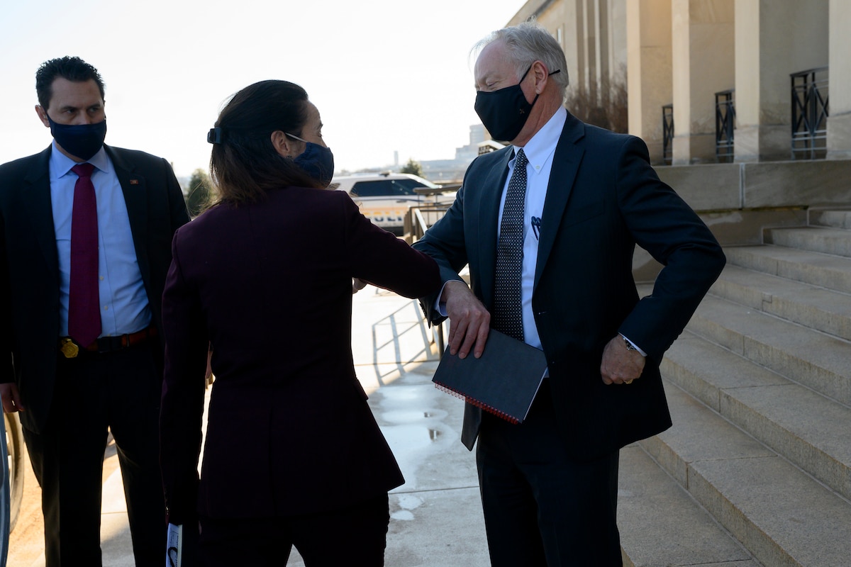 A woman and a man, both wearing face masks, touch elbows to greet each other while standing on steps outside a building.