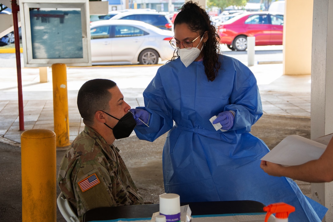 A medical technician, who’s wearing a face mask, gloves and gown, holds a swab up to a soldier’s nose.
