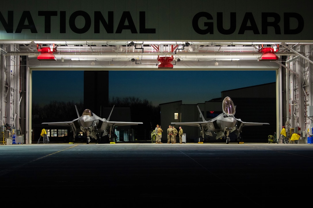 Service members stand between two jets near an open hangar door.