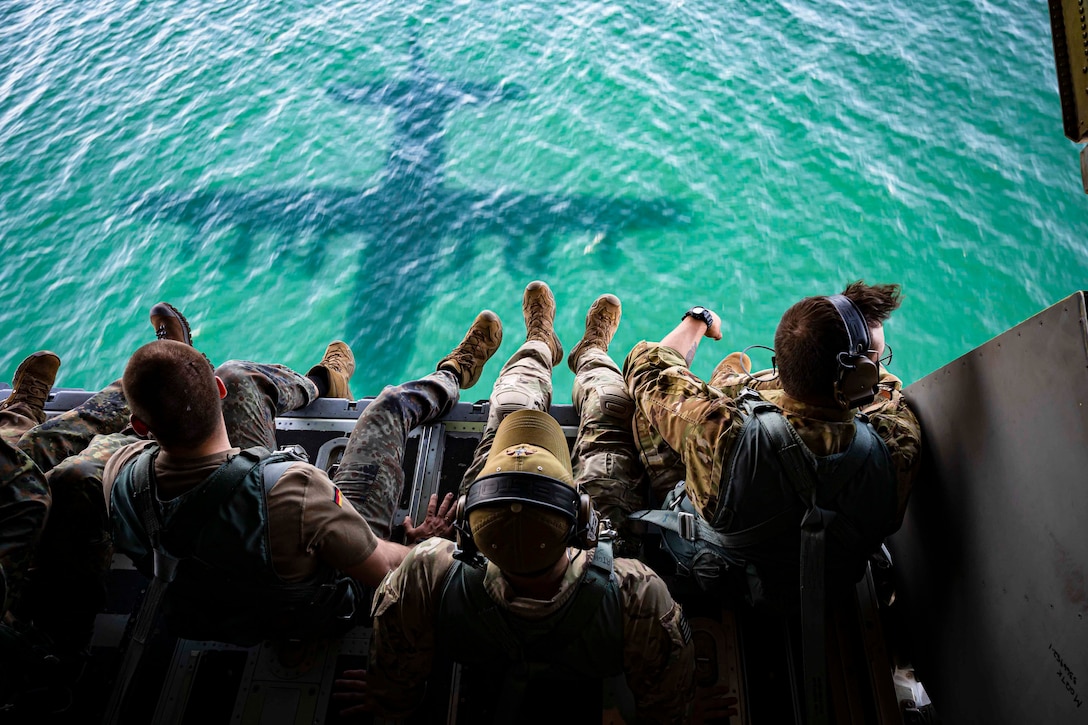 Airmen sit inside an airborne aircraft looking down at the aircraft’s shadow on the water.