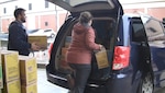 Two women and a man load cases of Girl Scout cookies into a blue mini-van at a warehouse loading dock.