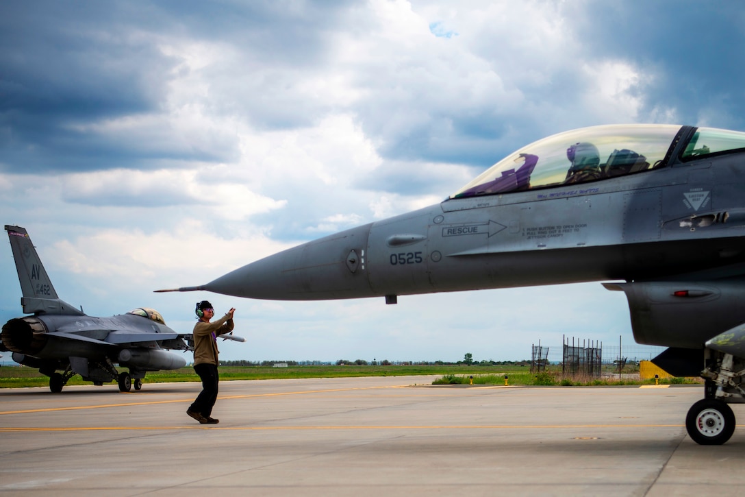 An airman on the ground guides another airman in a fighter jet.