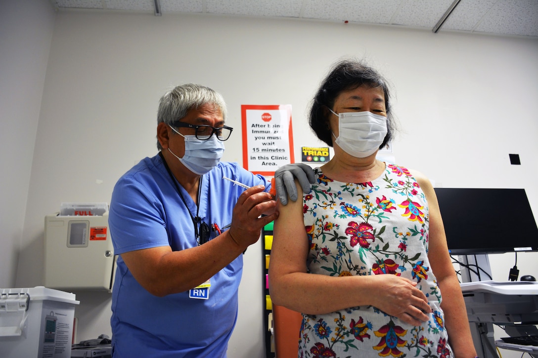 A medical tech wearing a face mask and gloves holds a syringe while administering a COVID-19 booster to a woman wearing a face mask.