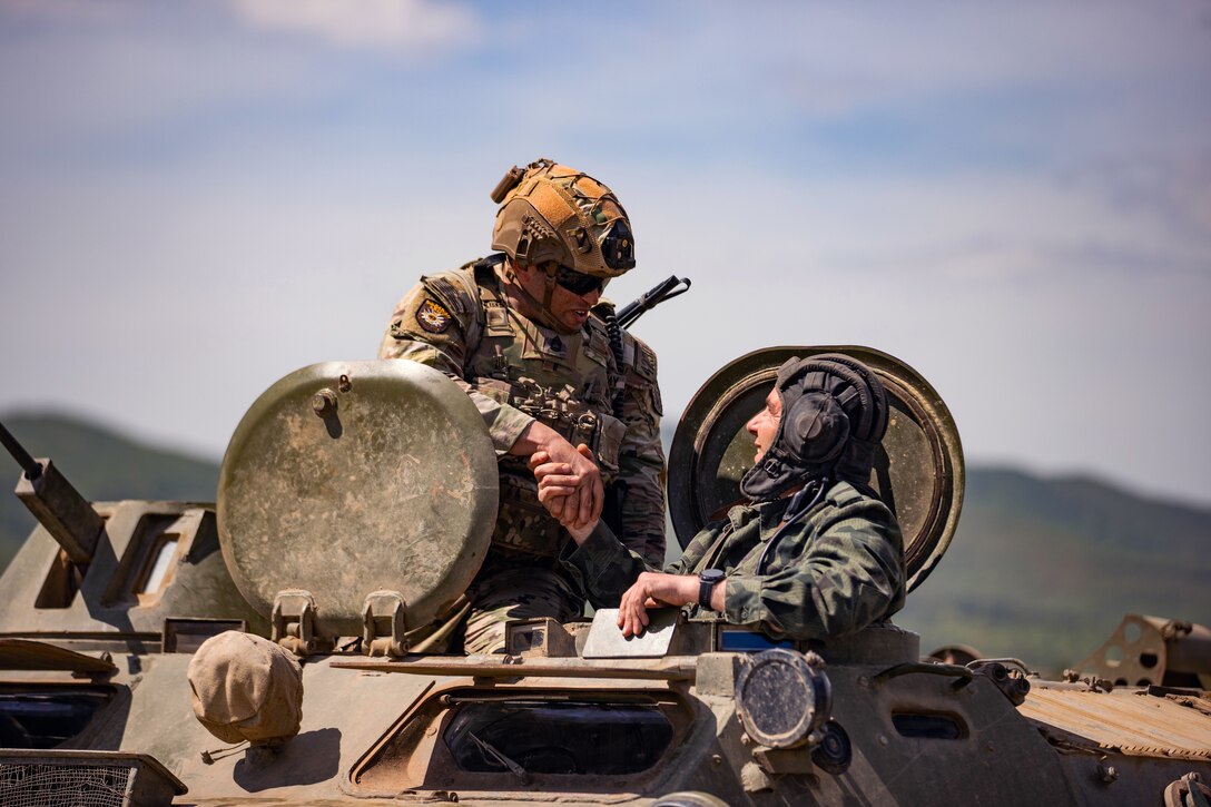 A soldier sits on top of a tank and shakes hands with a foreign military service member as he comes out of the tank.