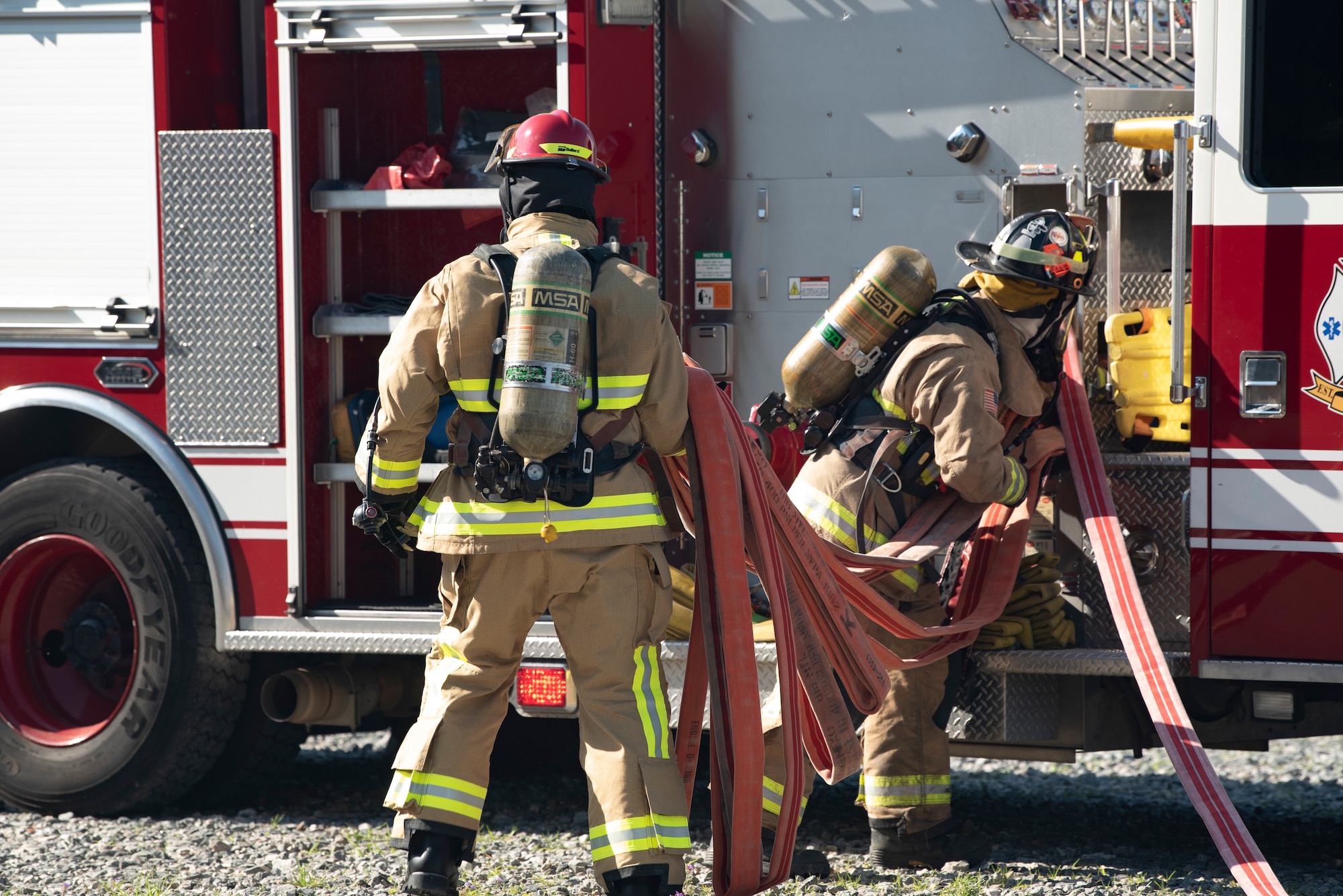 A photo of two members next to a fire stuck in fire fighter suits pulling a hose away from the fire truck.