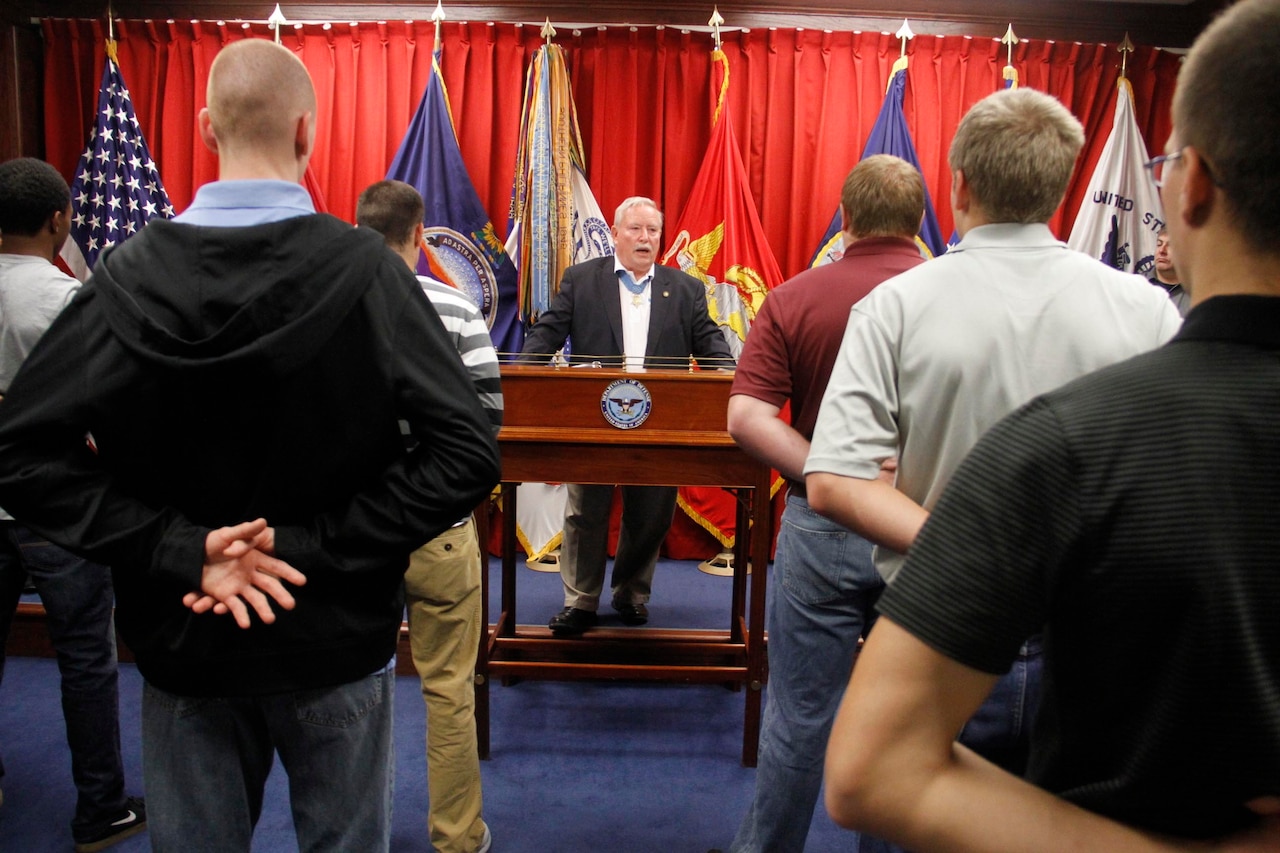 A man behind a lectern faces lines of men standing at parade rest in front of him.